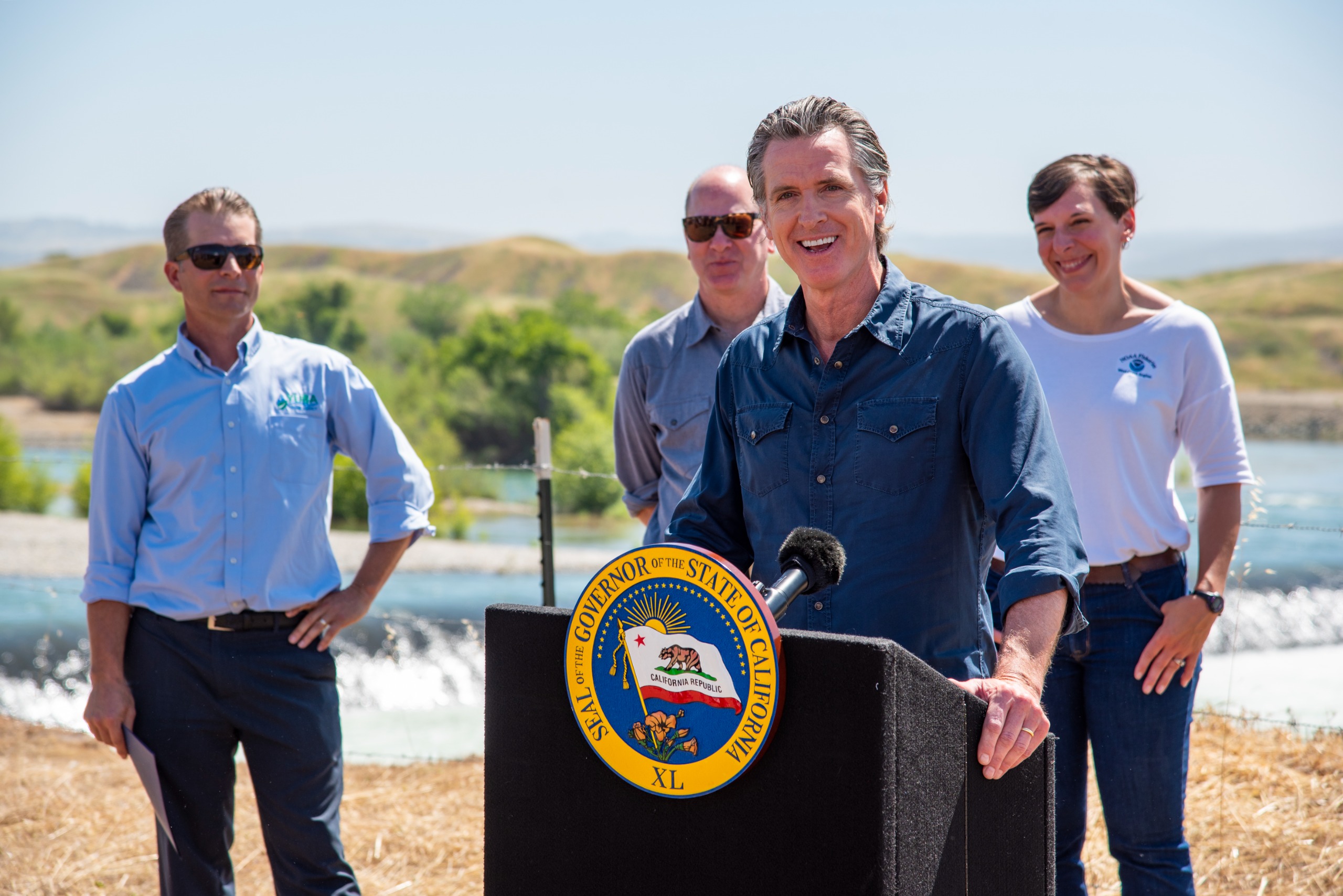 A man speaks at a podium with people and the lower Yuba River in the background. 
